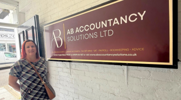 Angela Black, Director of AB Accountancy Solutions Ltd stands proudly next to her logo signage on the wall of her office.